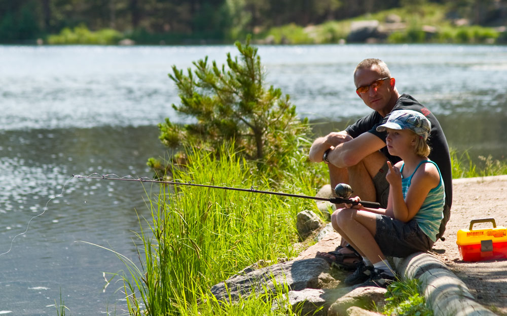 A parent and child sit at the edge of a lake in Estes Park, the child holding a fishing rod waiting for a bite 