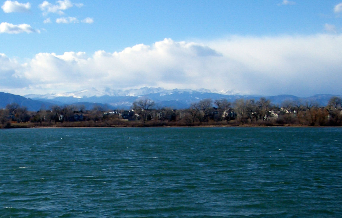 The blue-green water is rippling on Lafayette's Waneka Lake. In the distance, the snow-capped Front Range sits with blue skies and puffy white clouds.