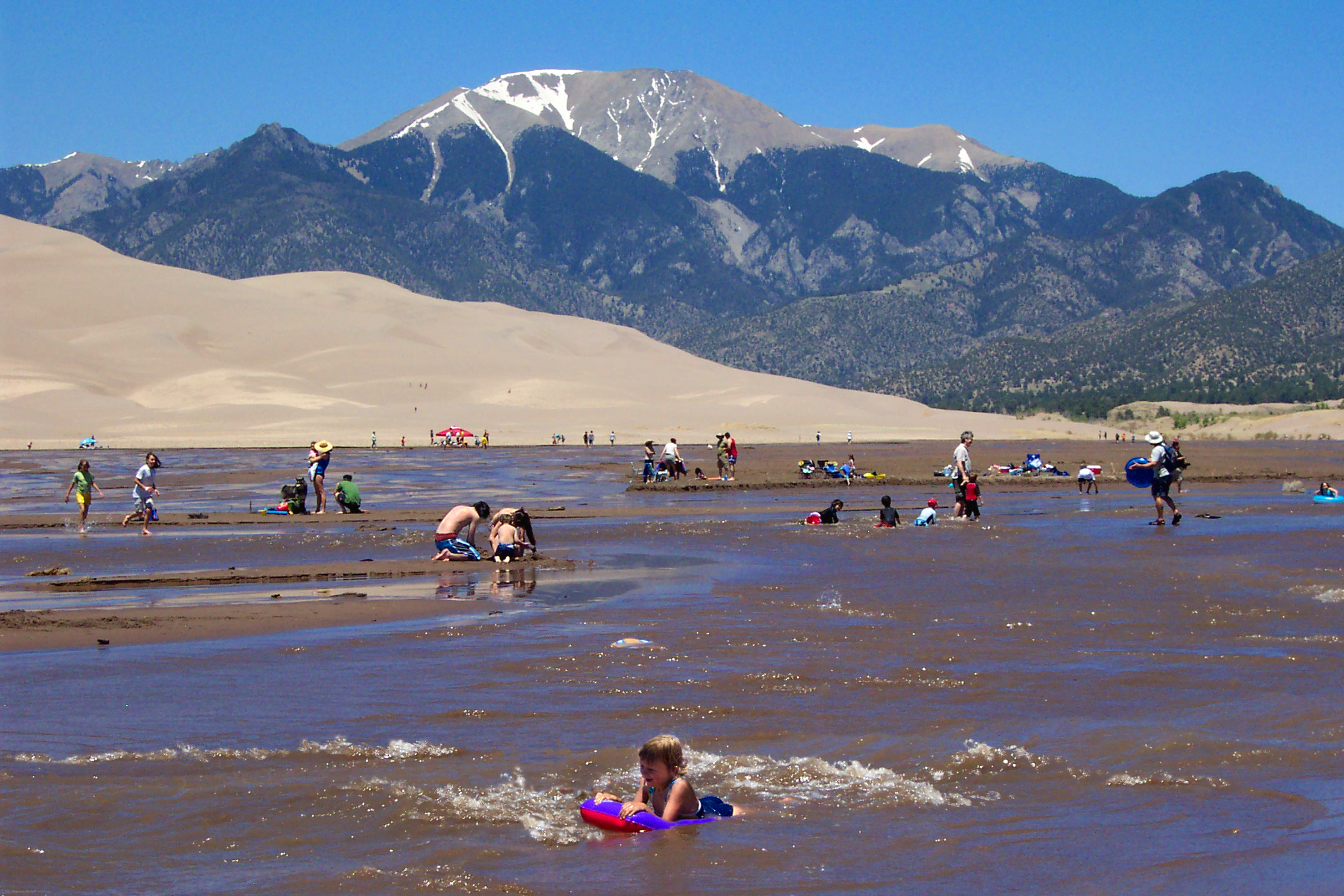 People play in the flowing waters of Medano Creek on front of the Great Sand Dunes. In the background Rocky Mountains soar into a blue sky with rivulets of white snow.