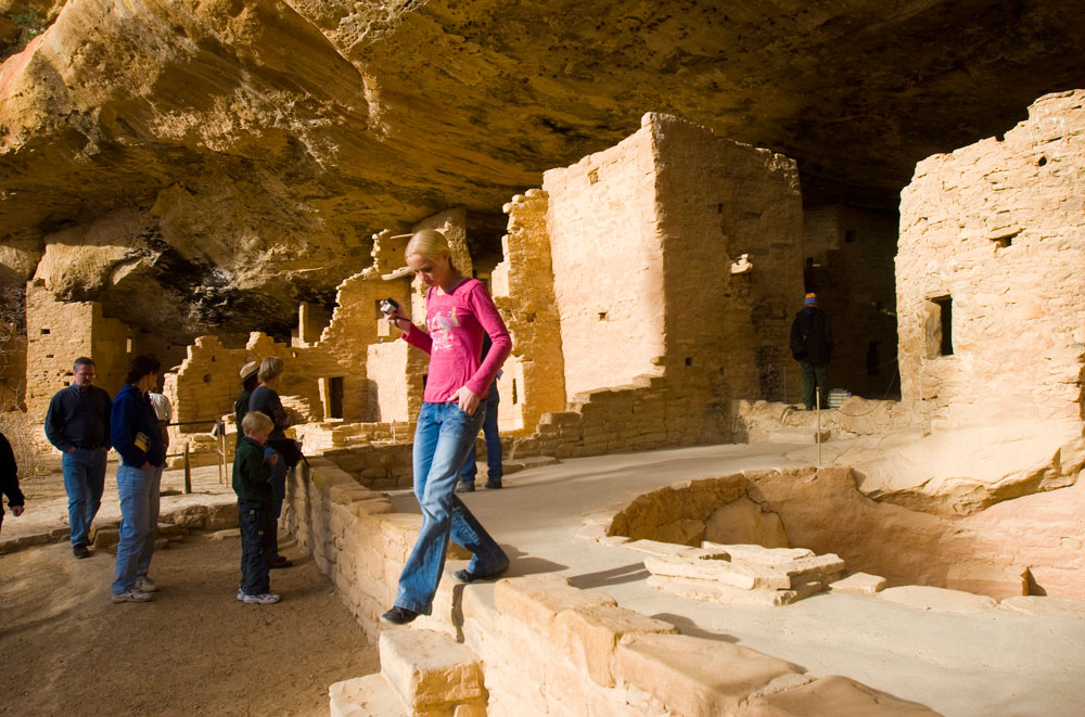 Someone in a pink shirt and jeans walks down steps under a tall beige cave at Mesa Verde National Park.