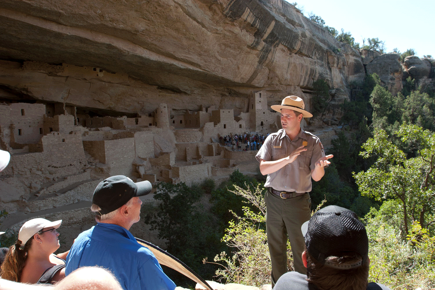 A park ranger speaks to a tour group at Mesa Verde National Park in Colorado. Behind the ranger, the cliff dwellings are shaded from the warm summer sun.