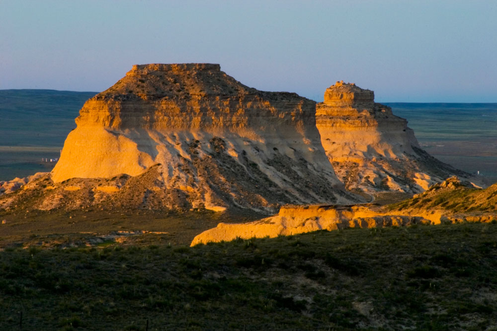Two rock formations rise out of the plains and tower of the surrounding Colorado landscape. The sunlight turns the rocks a soft yellow-orange color.