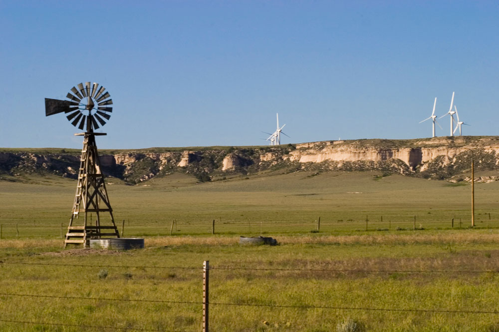 An old-fashioned windmill stands in a field at the Pawnee National Grassland in Colorado. On the other side of a rocky ridge, several modern, sleek air turbines spin.