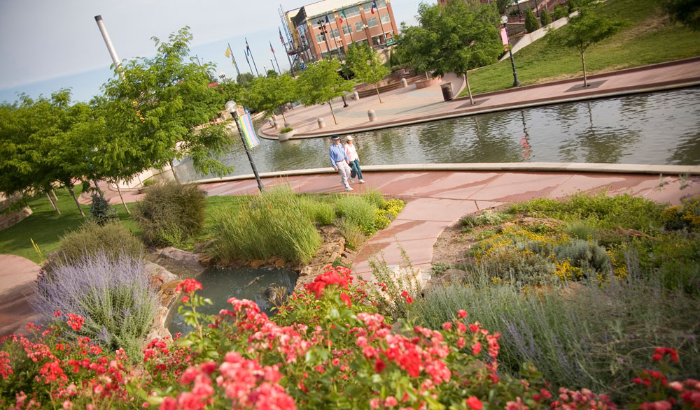 A couple walk hand in hand down a path of wide red-stone slabs that follows part of the Arkansas River in Pueblo, Colorado. Next to the path is a greenspace featuring young trees, lavender and red flowers.