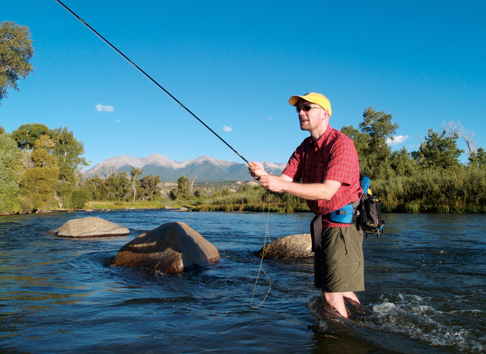 A flyfisher in shorts stands in knee-high waters of a blue river casting his line