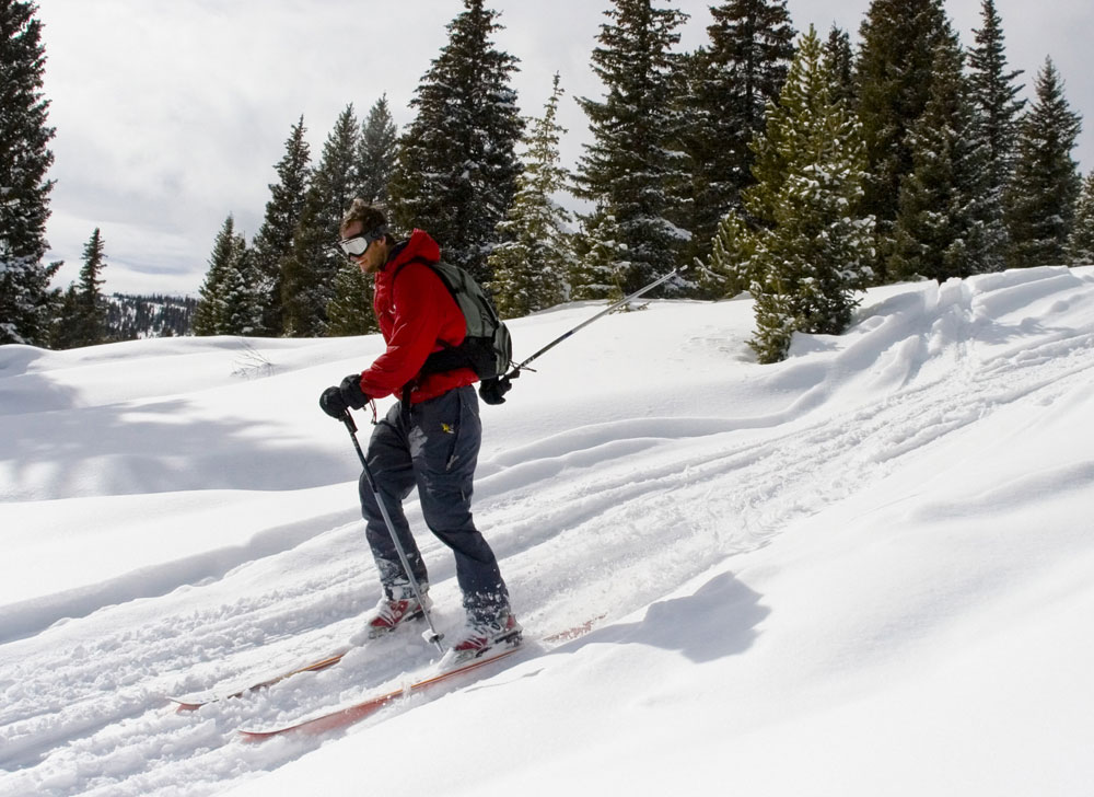 A male skier in a bright red jacket with black pants skis through Colorado's fresh and dry powder with goggles on a winter's day.