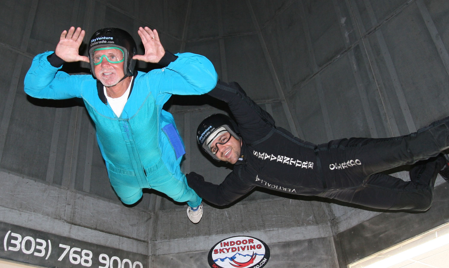 Two people, one in a teal fly suit and one in a black fly suit indoor skydive. 