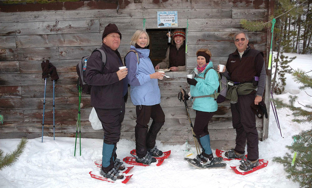 Four adults smile at the camera, all with hot chocolate in hand. They wear red snowshoes and appear to be taking a rest break at a wooden warming hut