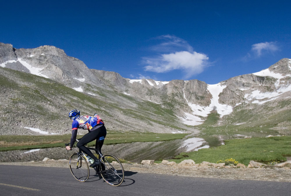 A cyclist zooms up a roadway under bright blue skies and a grassy meadow and a mountian ridge with a trace of snow