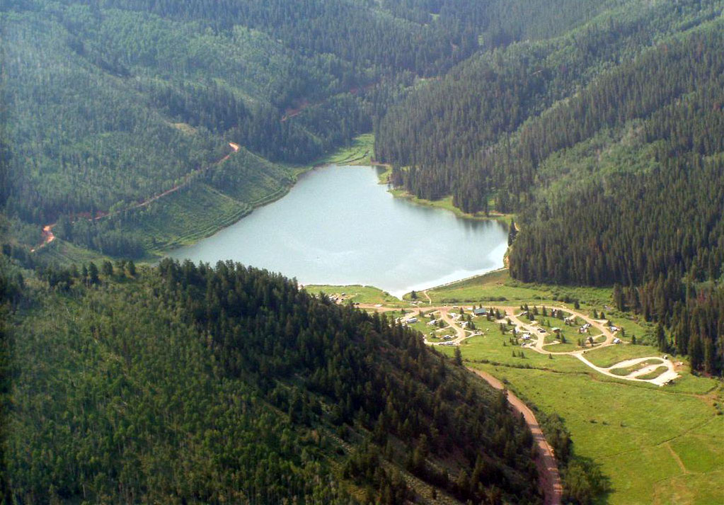 A lake is tucked into evergreen-tree covered mountains with a campground on green grass in the lower right.
