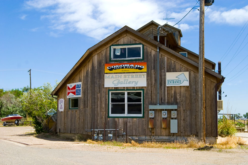 A wooden art gallery and feed store in Timnath on a blue-sky day. There are four signs on the building, on the right one has the Budweiser logo, in the middle a sunset colored sign says "Colorado Feed & Grain" and one below it says "Main Street Gallery." On the right a sign says "Entrance" with a blue arrow.