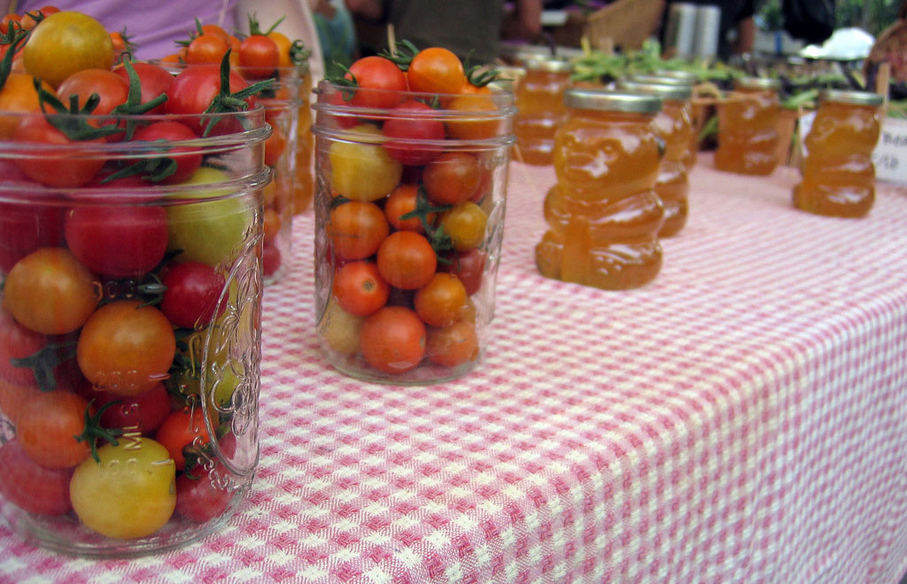 Yellow, orange and red cherry tomatoes sit in mason jars on a checkered tablecloth at a farmers' market in Boulder, Colorado. Bear-shaped jars on the table are filled with golden honey.
