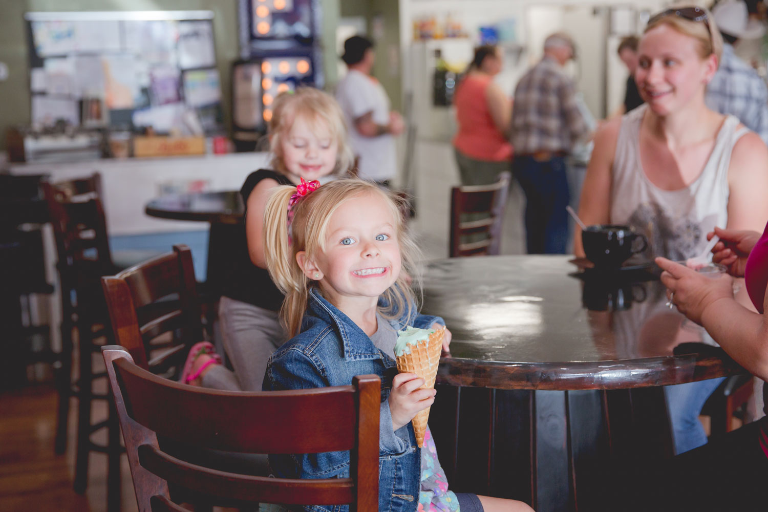 A child smiles at the camera while holding an ice cream cone as another child and woman look on. They're seated around a table and people are in the background.