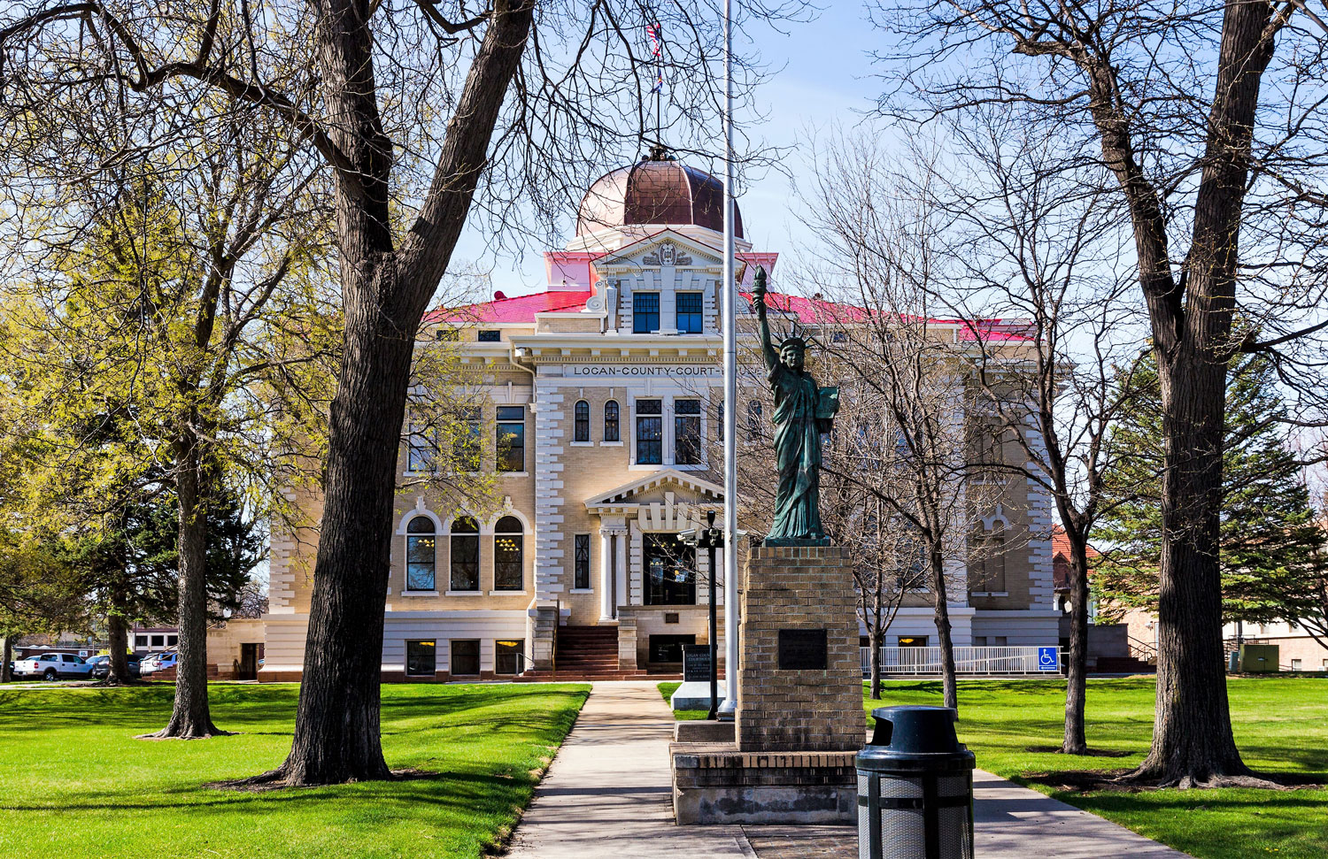 The Logan County Courthouse with a dome sits in the middle of a square with green grass and trees with whispy branches. In front of the flag pole, a small Statue of Liberty sits atop a brick pedestal.