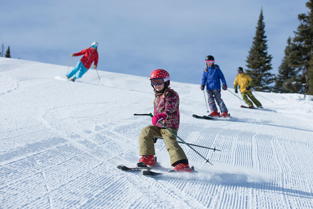 Kids skiing down a snowy mountain with pine trees in the background